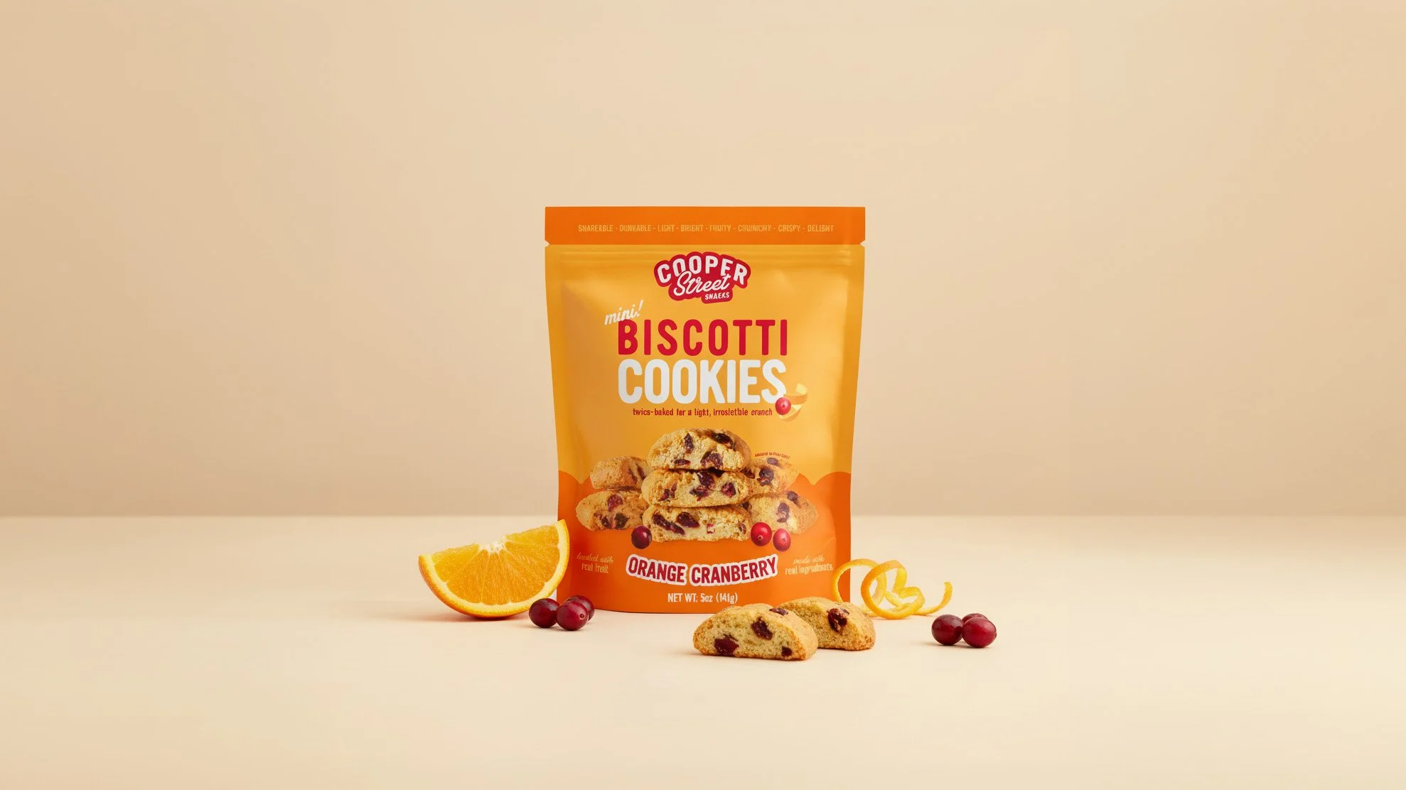 A bag of Cooper Street mini biscotti cookies with orange cranberry flavor on a plain surface, accompanied by an orange wedge, cranberries, and a few biscotti pieces in front.