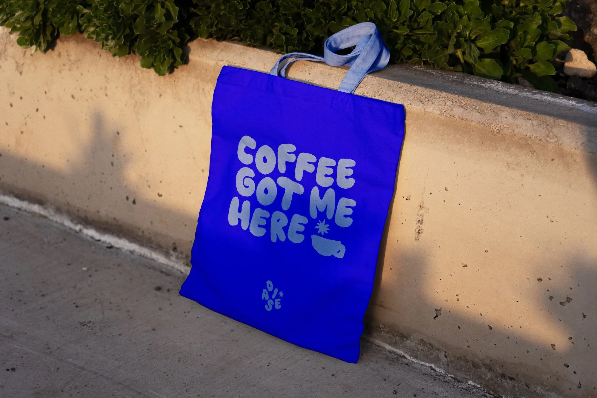 Blue tote bag with light blue handles hanging on a concrete ledge. The bag has the text 'Coffee Got Me Here' in playful bubble letters and a small illustration of a coffee cup.