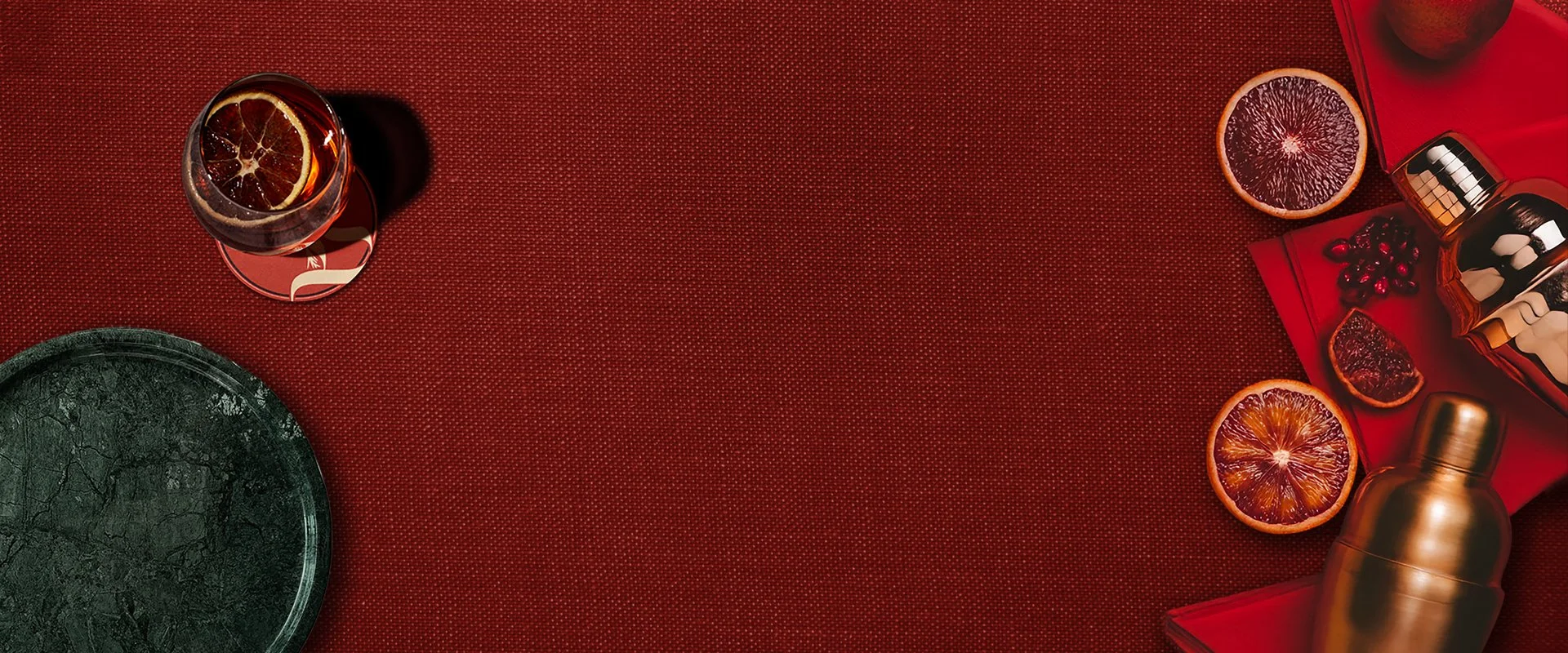 Flat lay of a green marble plate, a glass with a lemon slice, and sliced blood oranges on a red textured surface with red napkin, berries, and a golden cocktail shaker.