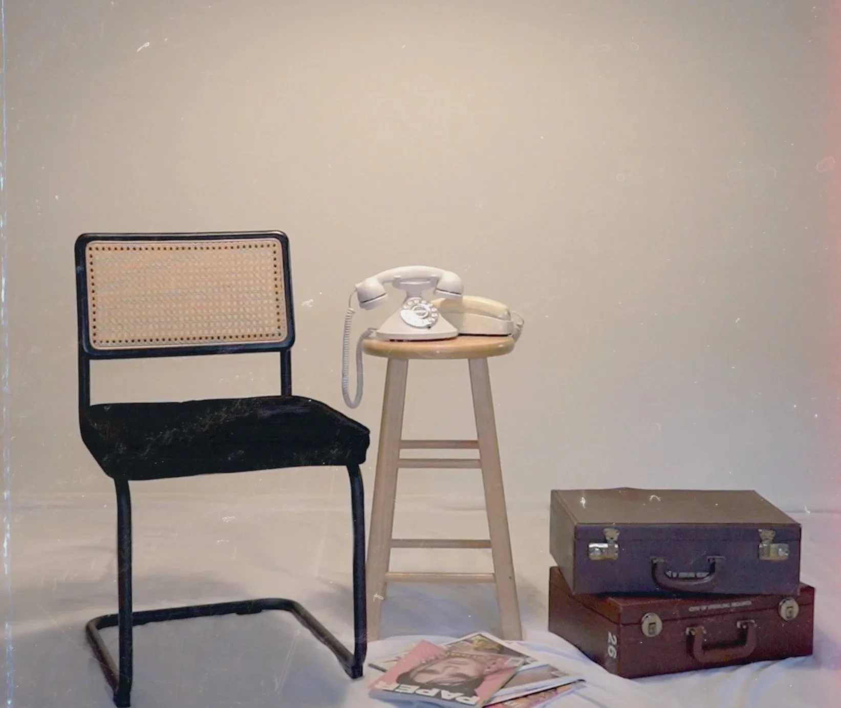 A black chair with a woven cane backrest, a wooden stool with an off-white rotary phone, a stack of three vintage suitcases, and a few magazines on the floor in front of a plain wall.