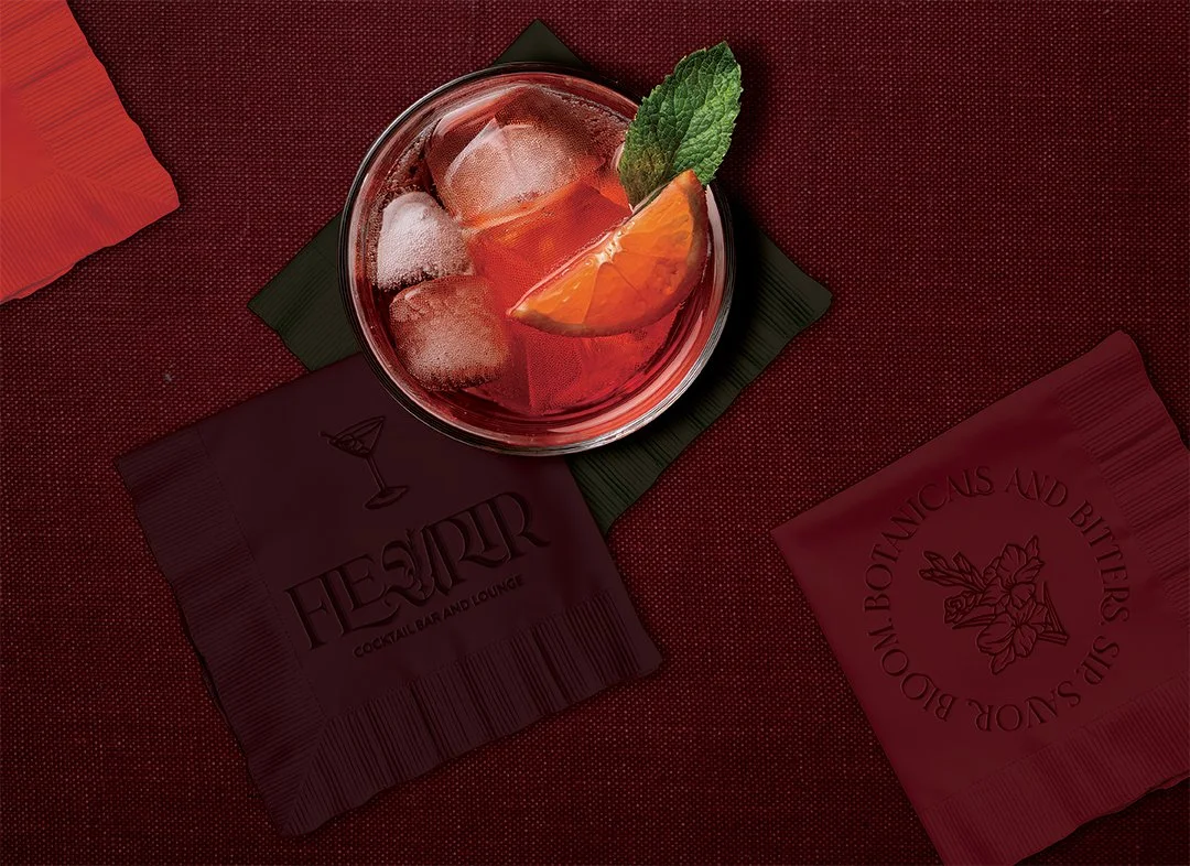 A cocktail with ice, an orange wedge, and a mint leaf in a glass, placed on a dark green napkin on a red textured surface, with two red embossed napkins.