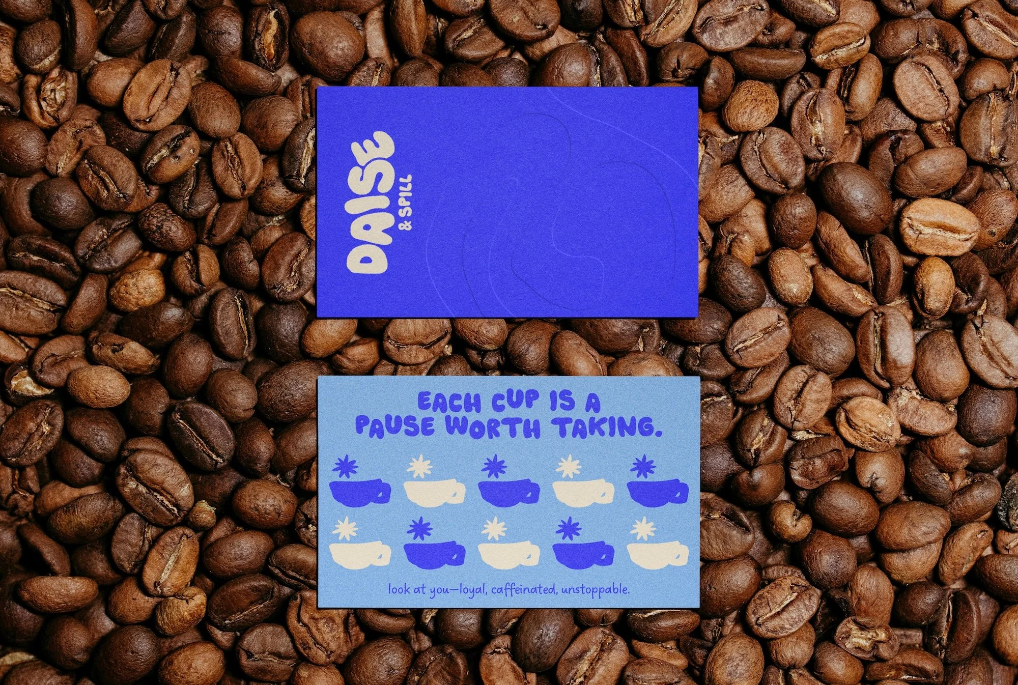 Two coffee cup cards placed on a bed of coffee beans. The top card is purple with the DAISE & SPILL logo. The bottom card has a blue background with the text "Each cup is a pause worth taking" and illustrations of coffee cups with stars and the phrase "look at you—loyal, caffeinated, unstoppable."