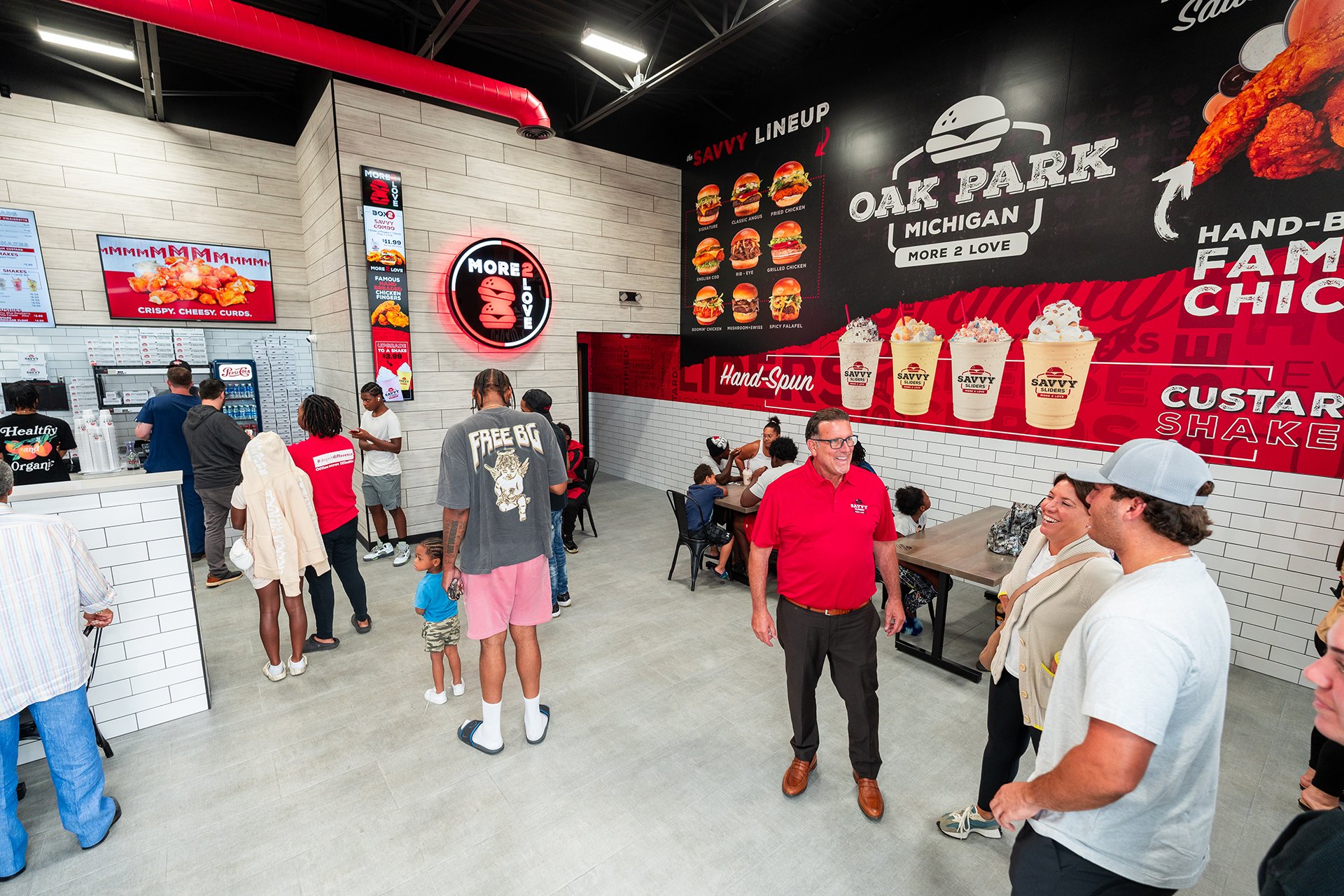 People inside a fast-food restaurant with menu boards, a counter, and a large signage for Oak Park, Michigan, loaded with images of burgers, shakes, and chicken dishes.
