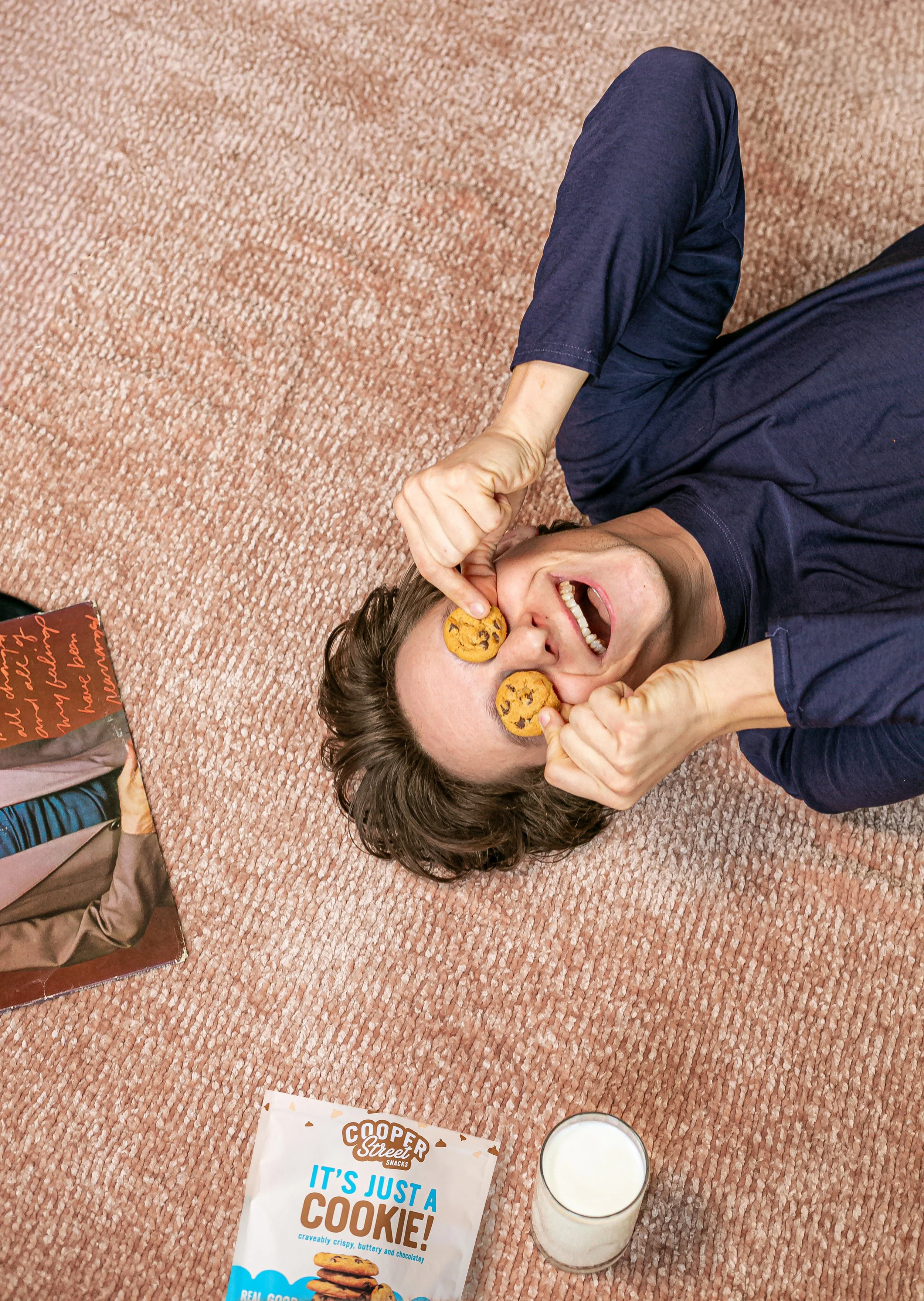 A man lying on a pink carpet, smiling and holding two chocolate chip cookies to his eyes while pretending they are eyes, with a glass of milk, a cookie package, and a cookie book nearby.