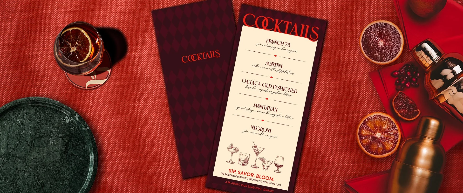 A bar or restaurant table with a red textured tablecloth, a cocktail menu, a glass with a citrus slice, and various citrus slices, a garnished cocktail shaker, and decorative items.