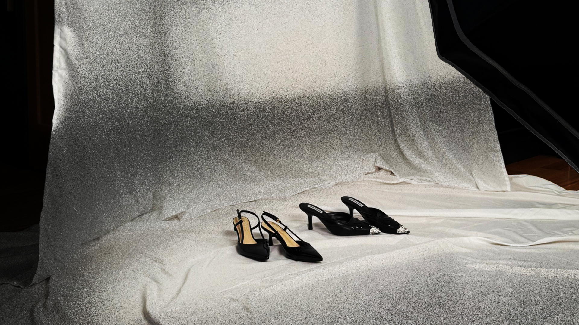 Two pairs of black high-heeled shoes on a light-colored fabric surface, with a backdrop of similar fabric, possibly in a photography studio setup.