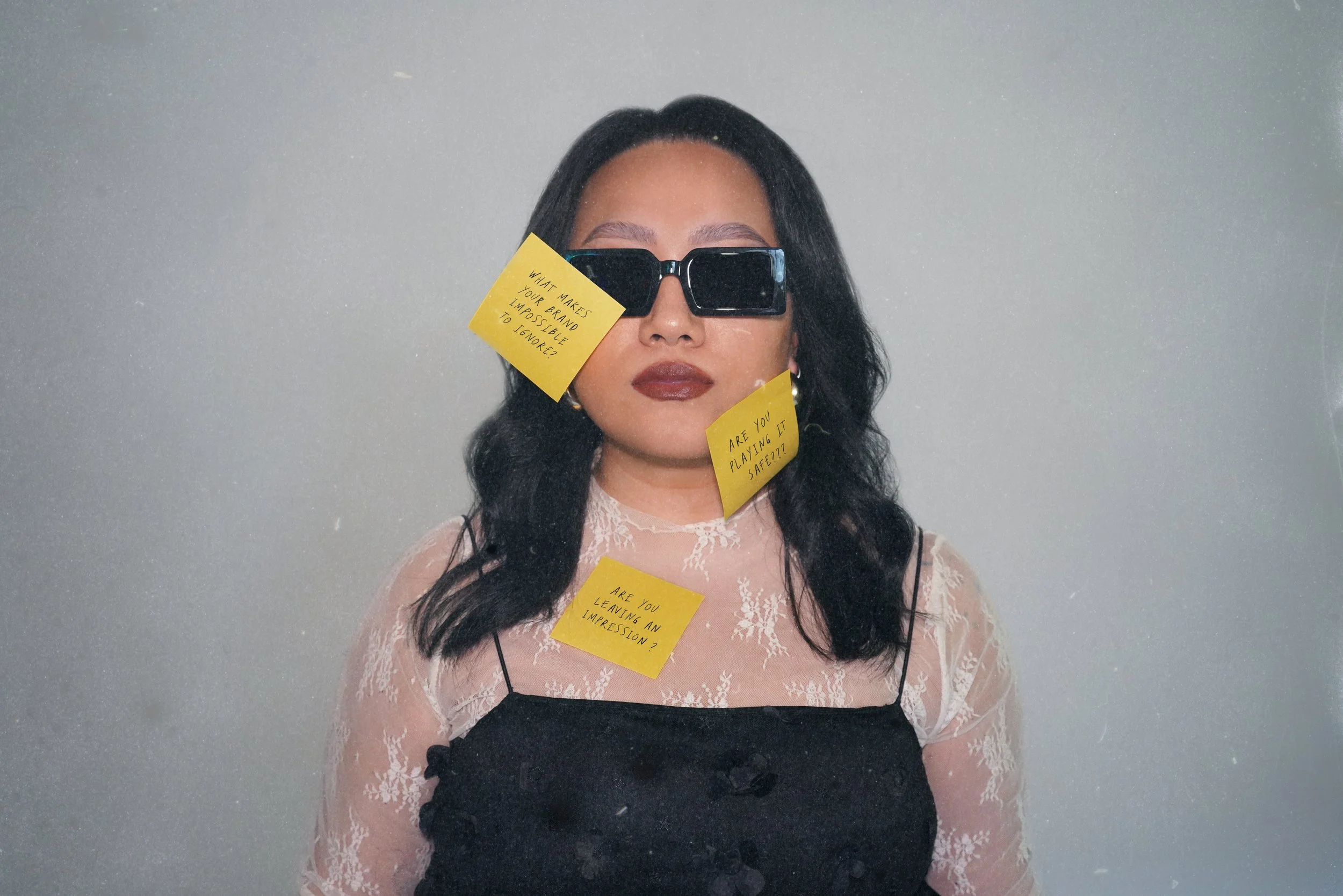Woman with black hair, wearing sunglasses and a lace top, has three yellow sticky notes on her face and chest with handwritten questions.