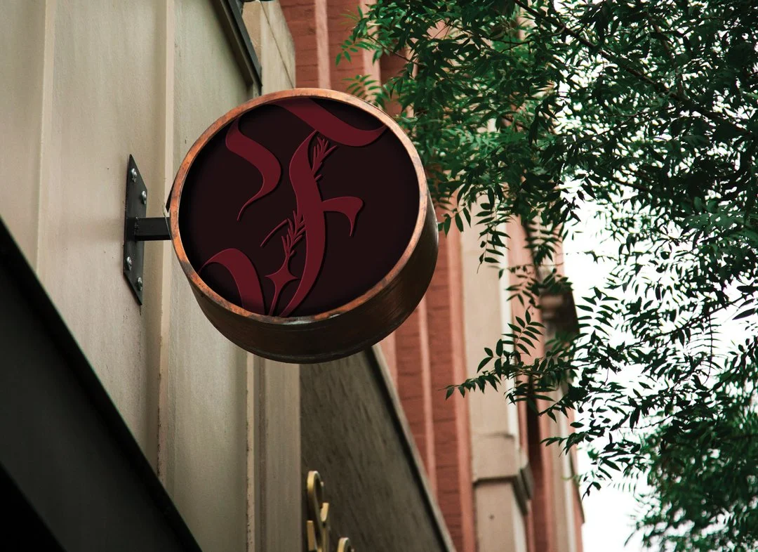 A circular sign with a dark background and a stylized 'F' for 'Fleurir' in red, attached to the side of a building, with green foliage nearby.