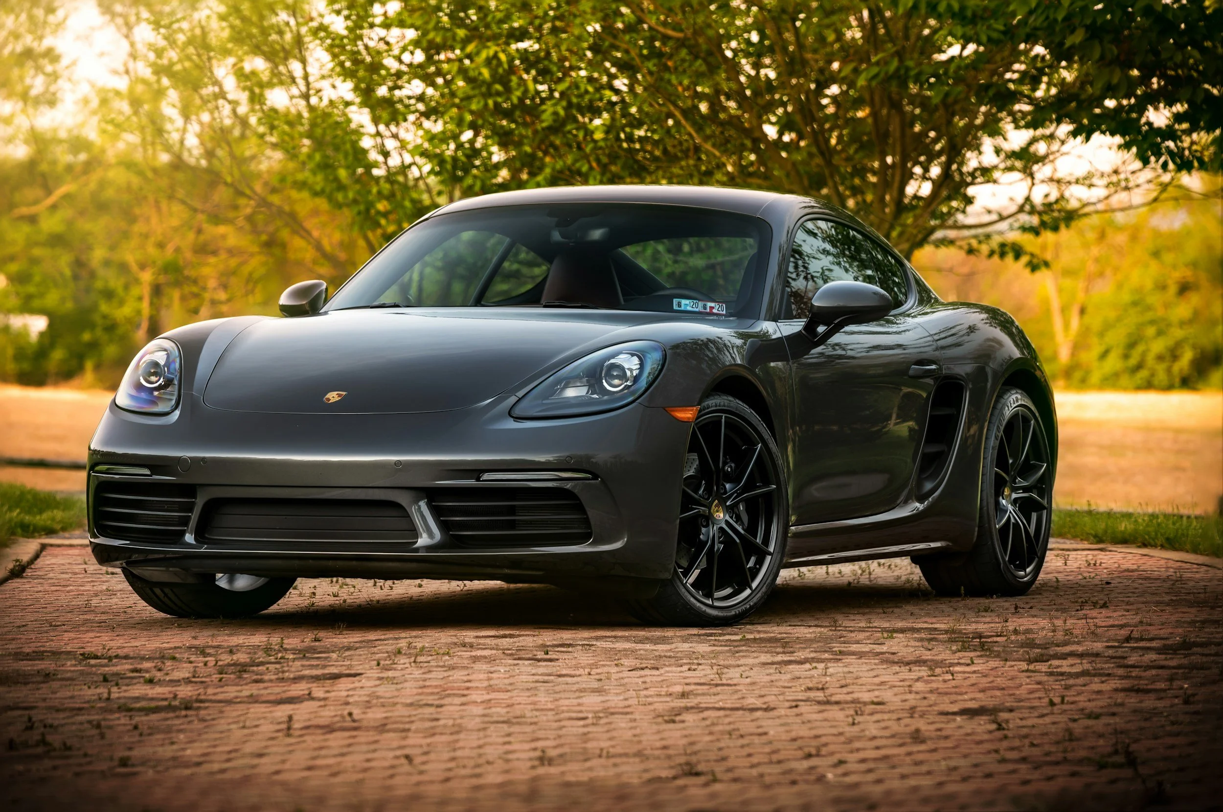 Black Porsche sports car parked on a brick driveway surrounded by trees with green foliage in the background.
