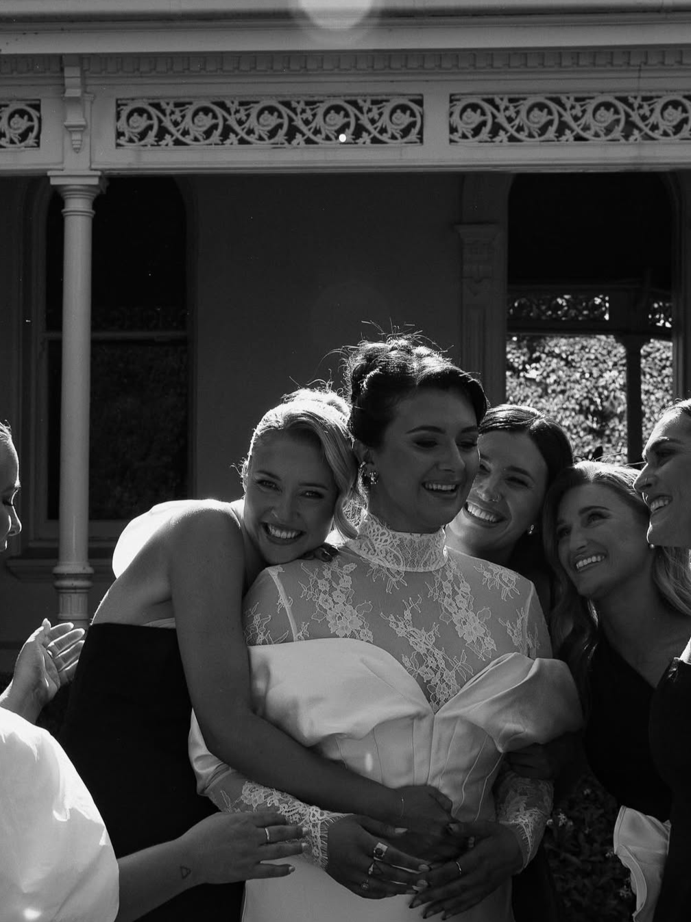 A bride in a lace wedding dress smiling with five bridesmaids hugging her outside.