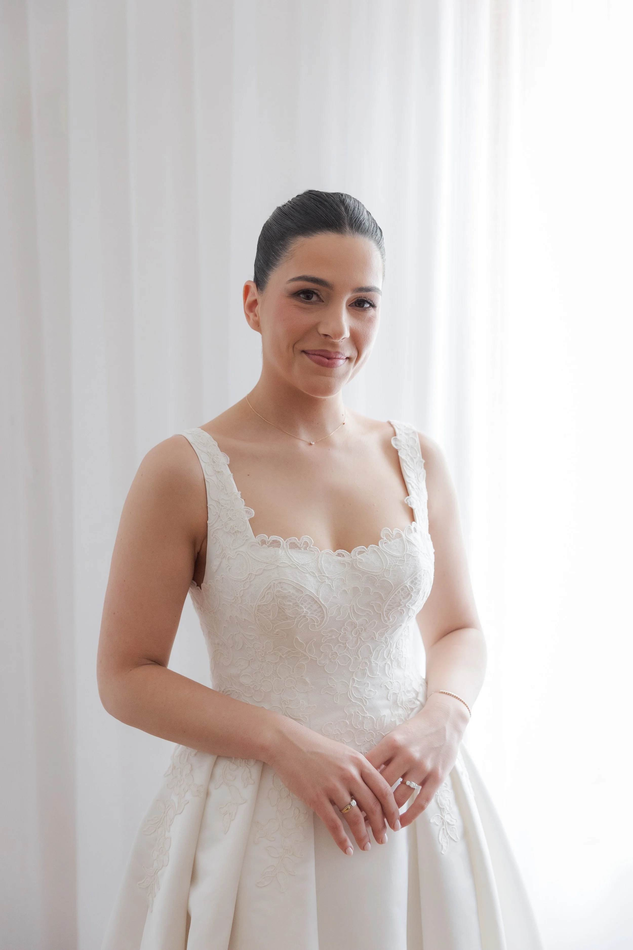 A woman in an elegant wedding dress with lace details, standing in front of white curtains, smiling softly at the camera.