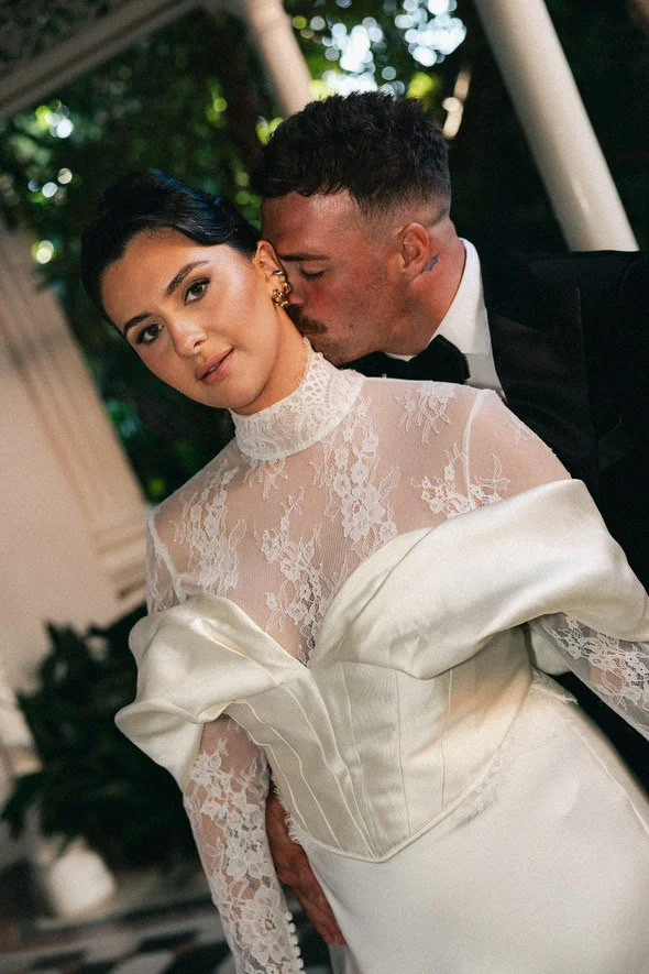 A woman in a white lace wedding dress with a high neckline and puffy sleeves is being kissed on the cheek by a man in a black tuxedo. They are outdoors with blurred greenery in the background.