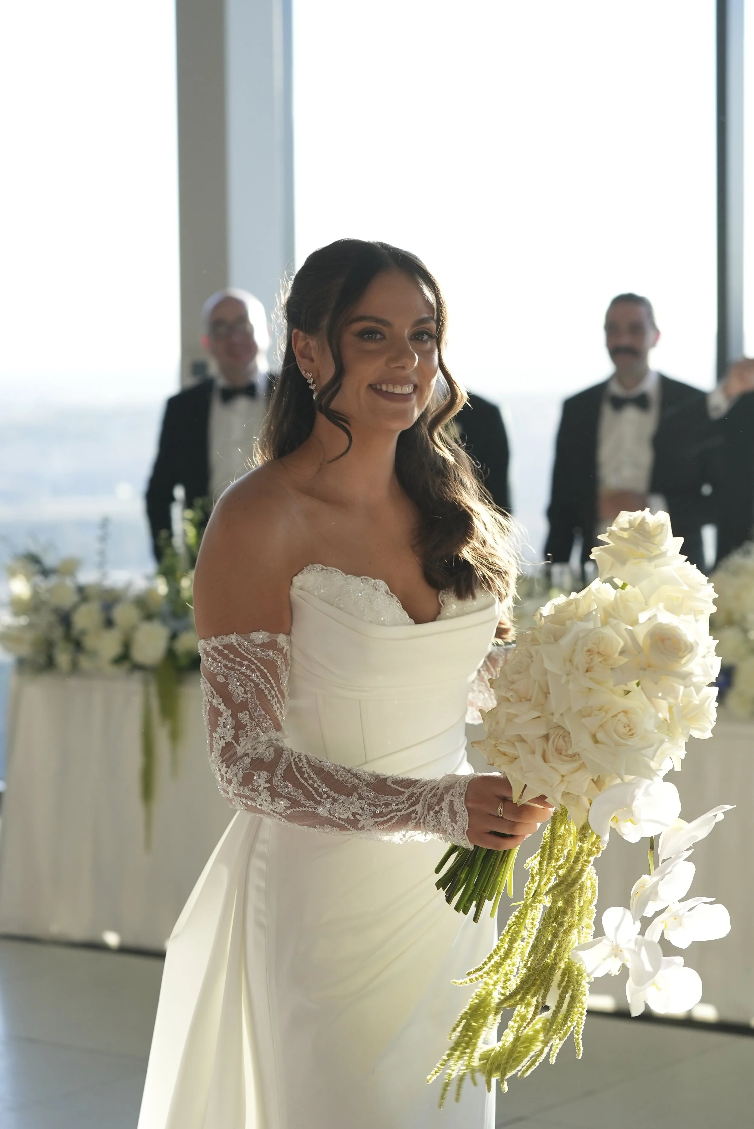 Bride smiling in a white wedding dress holding a bouquet of white roses, with three men in tuxedos in the background, in a bright, modern venue with large windows.