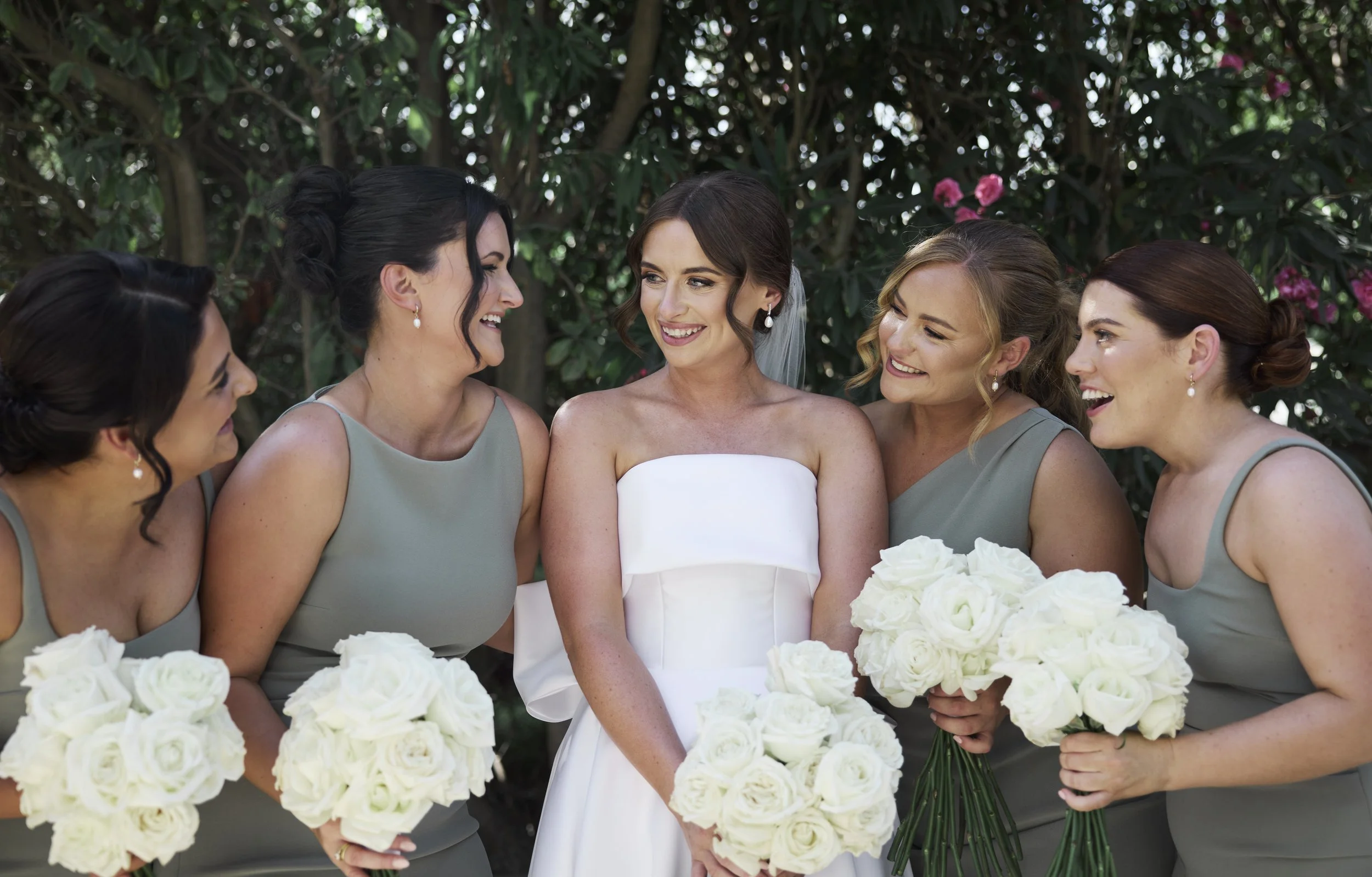 A bride in a white strapless wedding dress surrounded by four bridesmaids in gray dresses, all holding white bouquets, standing outdoors in front of green foliage and pink flowers.