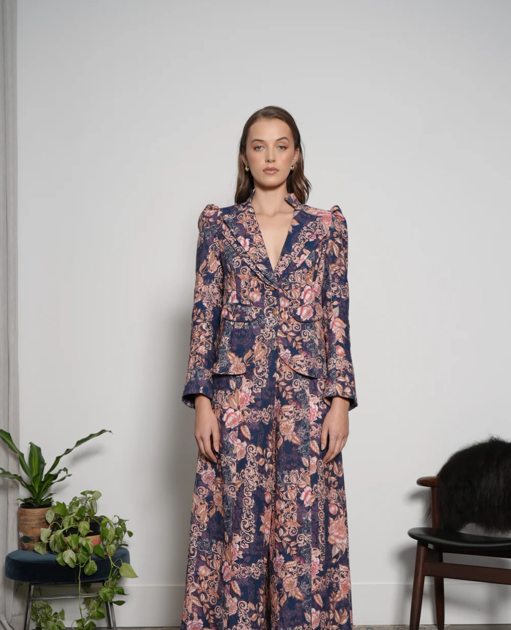 A woman with brown hair wearing a patterned suit with a blazer and wide-leg pants standing against a plain white wall.