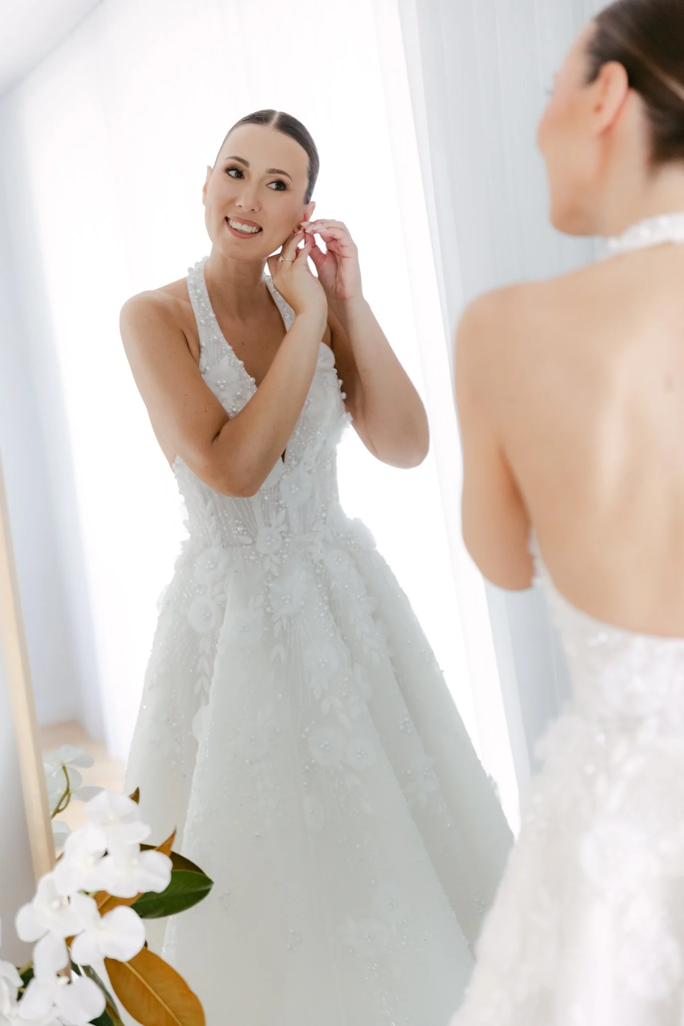 A bride in a white wedding dress is adjusting her earring while looking in the mirror.