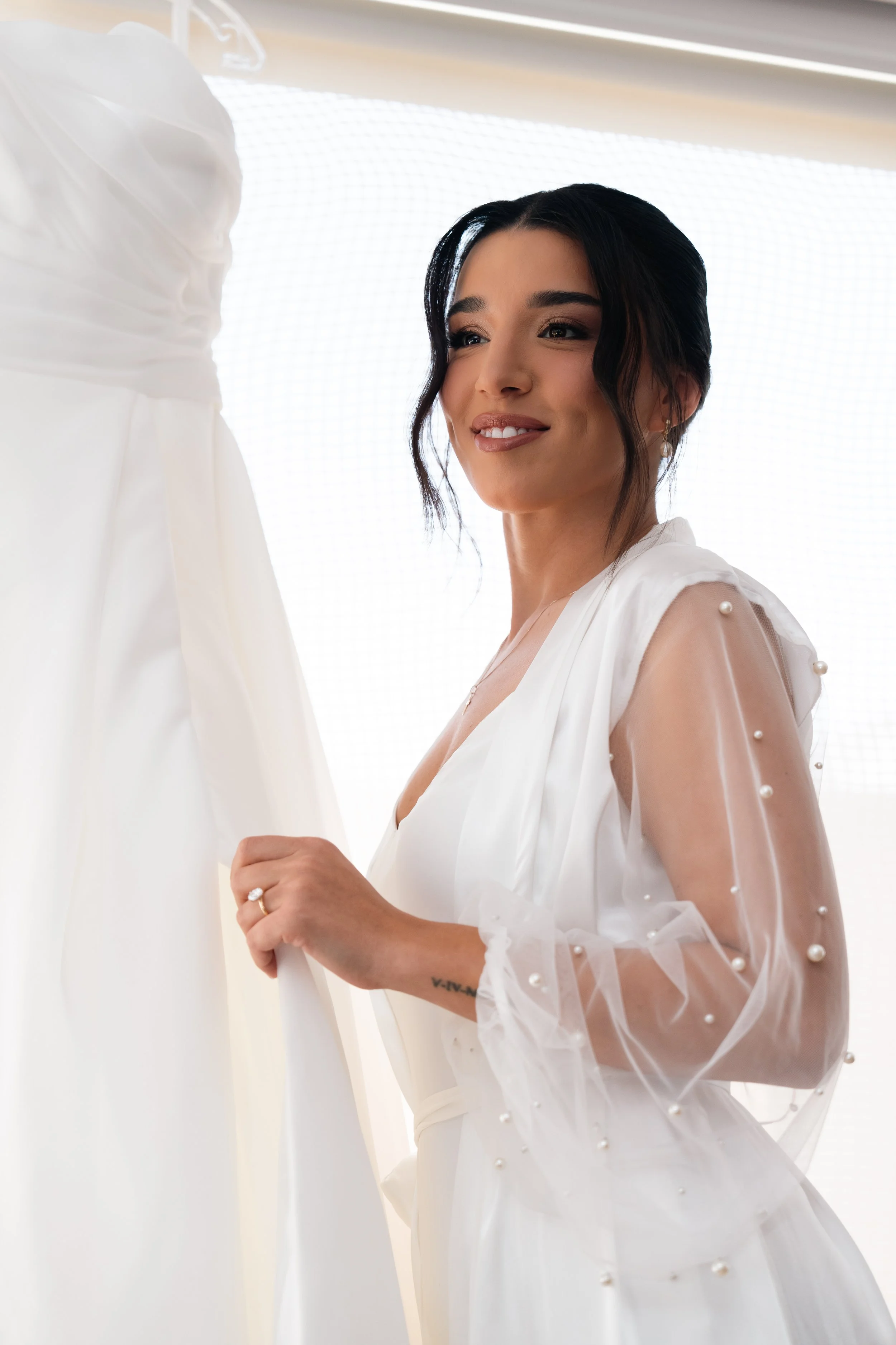 A woman in a white dress with sheer sleeves decorated with pearls, standing next to a white dress on a hanger, smiling.