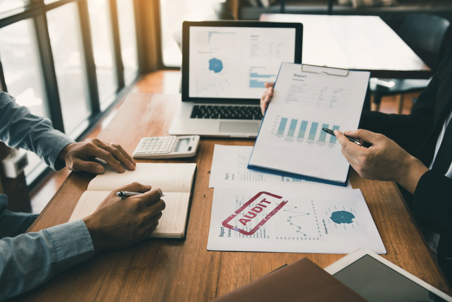 Business meeting with two people reviewing an audit of financial reports and documents on a wooden table, with a laptop showing charts and a tablet nearby.