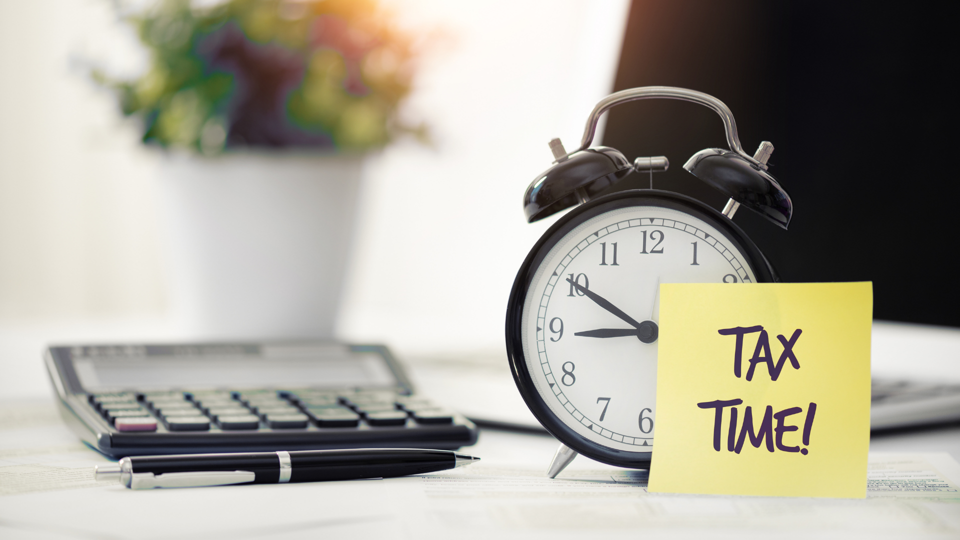 An analog alarm clock showing 9:47, with a yellow sticky note that says 'TAX TIME!' in black letters, a calculator, a pen, and a blurred computer keyboard and plant in the background on a desk.