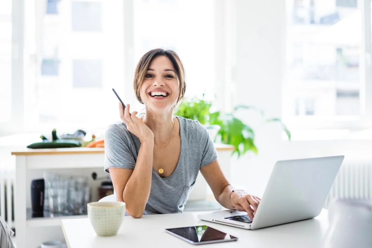 A woman sitting at a table with a laptop, smartphone, and mug, smiling and holding a pen near her face in a bright, modern kitchen.