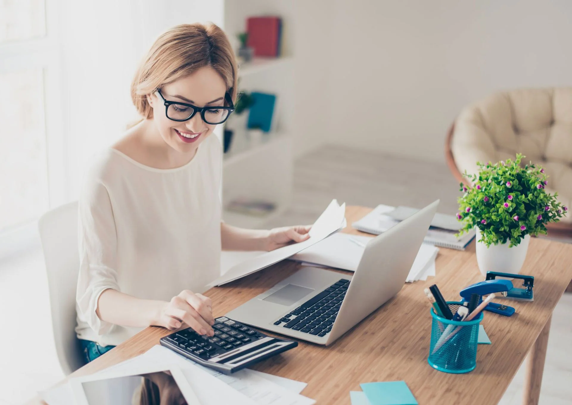 Smiling woman with glasses working at a desk with a laptop, calculator, papers, and office supplies, in a bright, home office setting.