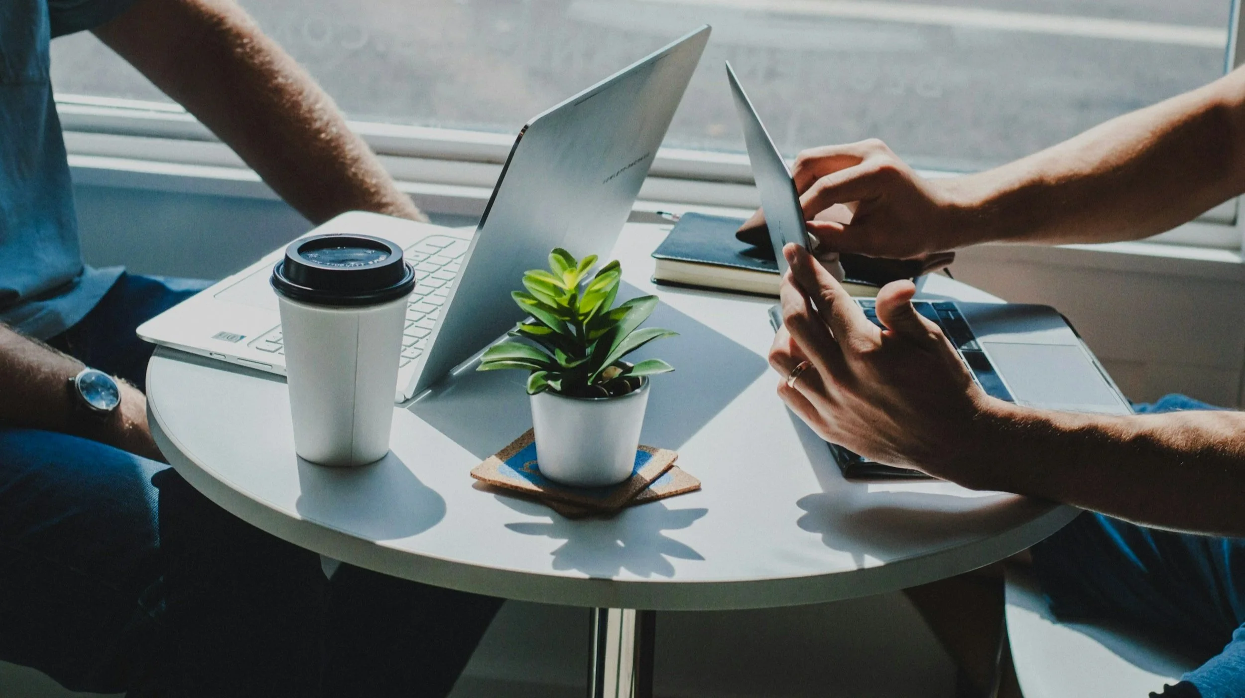 Two people working at a round table with a laptop, a tablet, a small potted plant, a takeaway coffee cup, a closed notebook, and a smartphone, near a window with natural light.