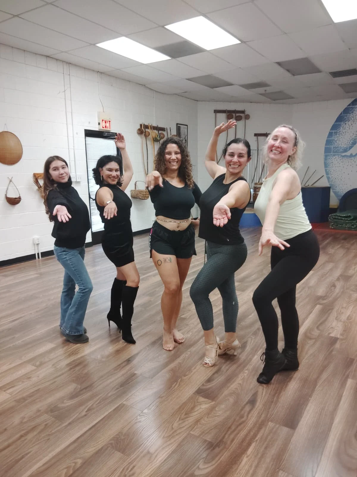 Five women in dance attire and workout clothes pose with arms raised and smiling in a dance studio with wooden floors and decor on the walls. Samba class in sacramento