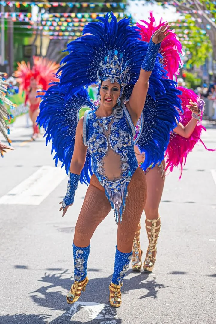 Woman in a blue and silver carnival costume with large feathered headdress, dancing during a parade, with other dancers in colorful costumes in the background.