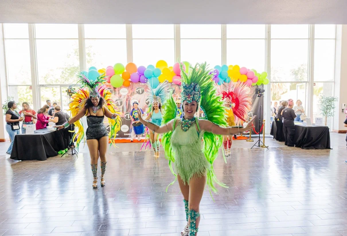 Women performing a samba dance in a brightly decorated event space with balloons and colorful banners in the background, observers on either side.