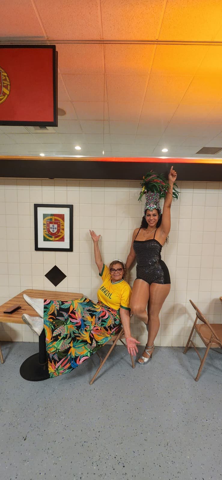 Two women posing in front of a Portuguese flag on a tiled wall. One woman is sitting on a chair, wearing a colorful skirt and a yellow Brazil shirt, smiling and raising her arm. The other woman is standing, wearing a sparkling black outfit and an ela