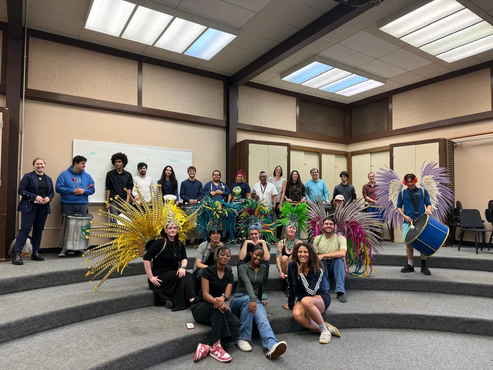 Group of people, some in colorful feathered costumes, standing and sitting on a stage in a room with a whiteboard and a patterned ceiling, posing for a photo.  G.R.E.S. unidos da capital of sacramento brazilian samba performing arts group