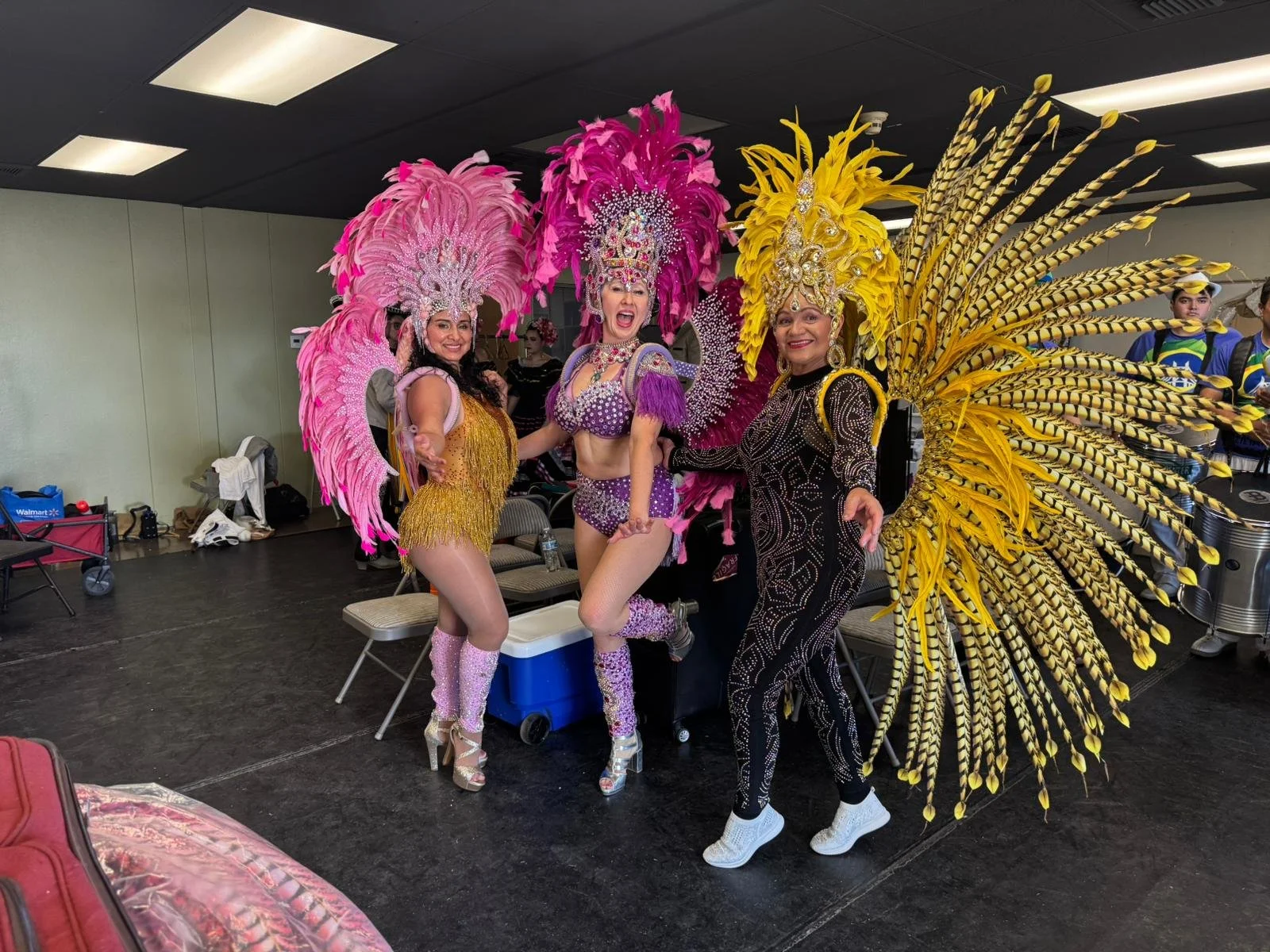 Three women in colorful, elaborate feathered costumes posing backstage at a carnival or parade, with chairs and miscellaneous items in the background.