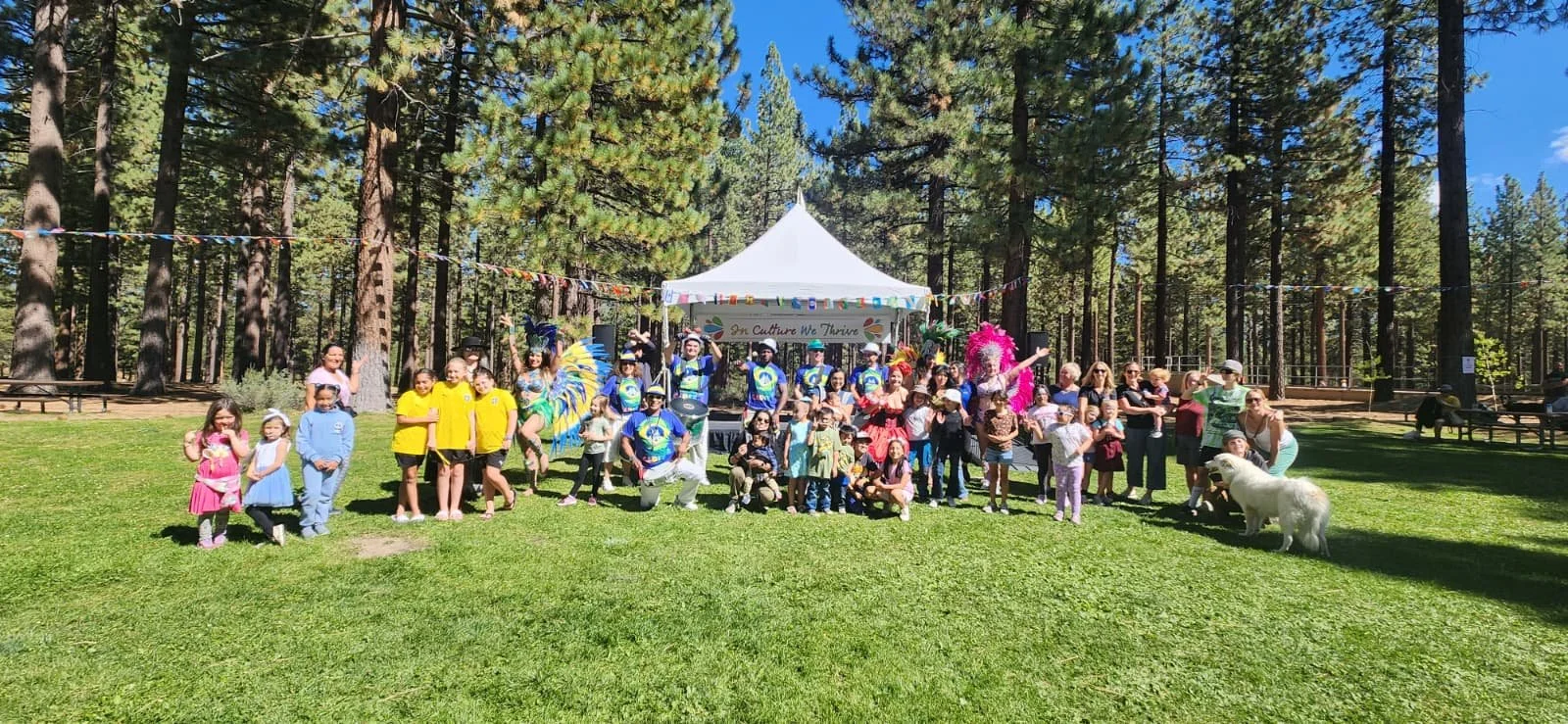 A large group of people, including children and adults, gathered outdoors on a grassy area surrounded by tall pine trees. There is a white event tent with a banner that reads 'In Culture We Thrive'. People are dressed in colorful clothing, some weari