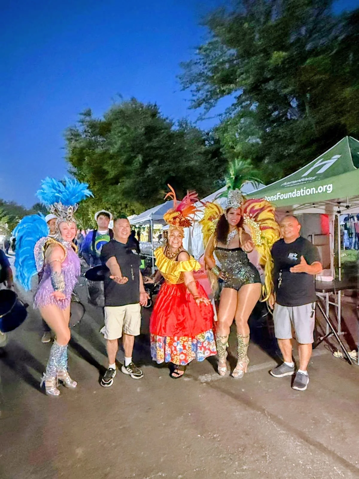 Group of six people, including women dressed in elaborate carnival costumes with feathers and sequins, posing outdoors at night with tents and trees in background.