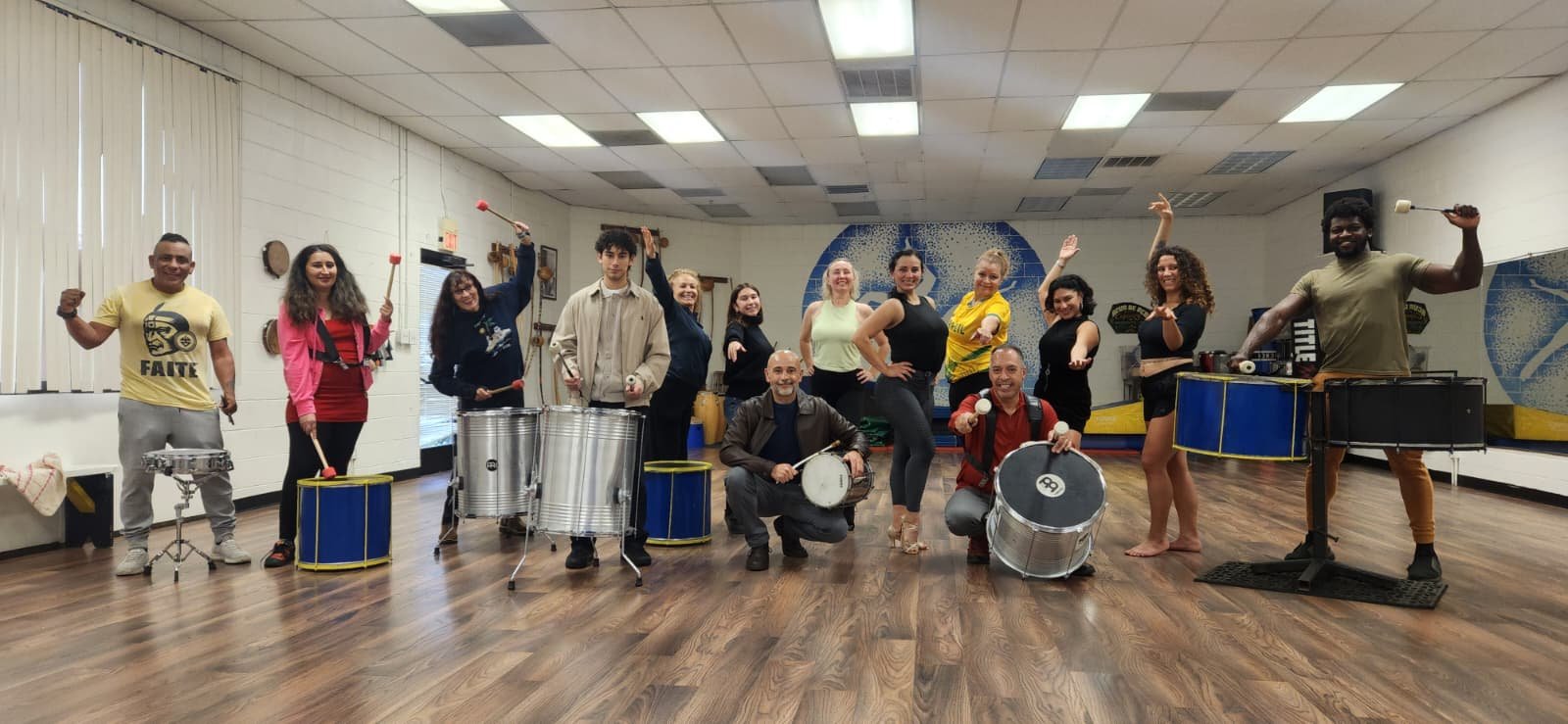 Group of twelve diverse people in a dance studio, holding percussion instruments, smiling and posing for a photo.