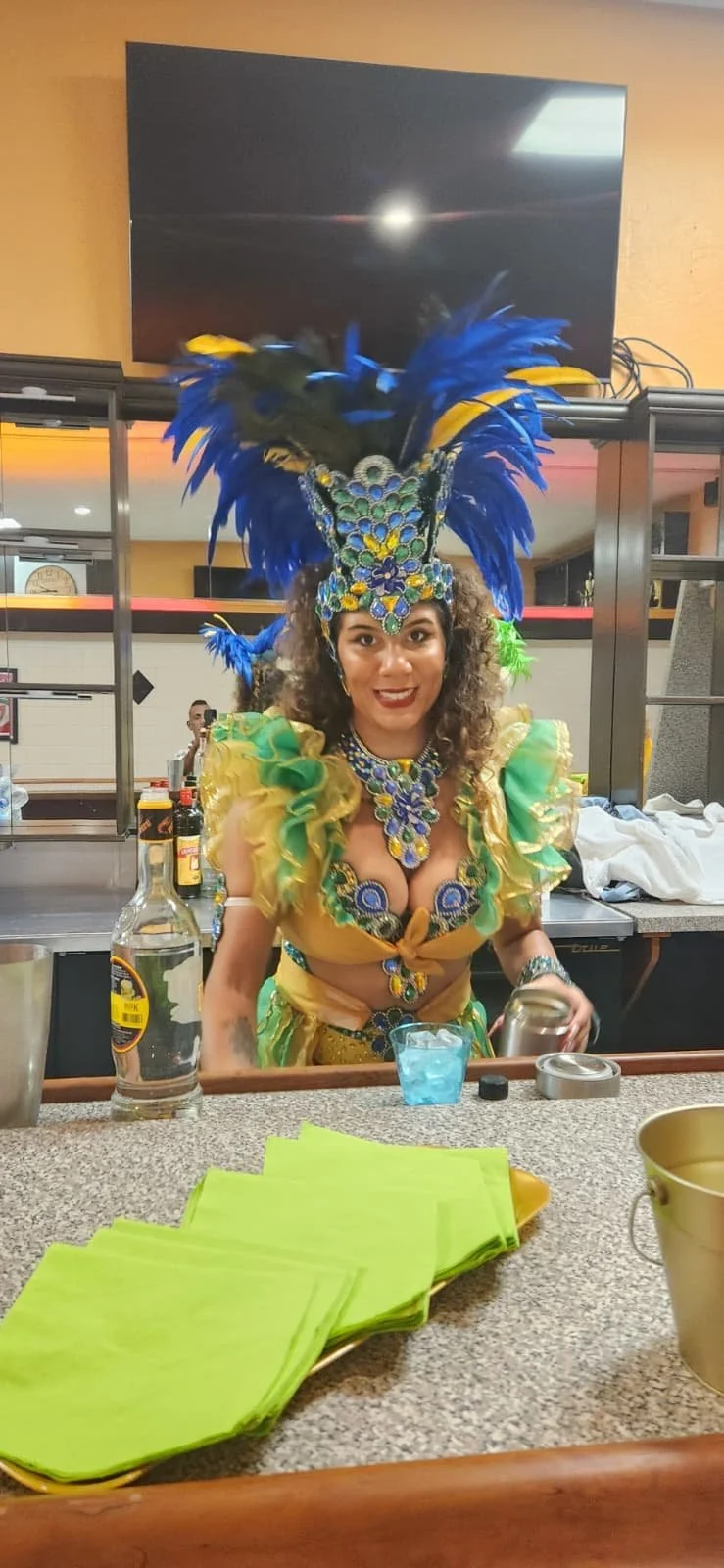 A woman dressed in a colorful carnival costume with a large feathered headdress, standing behind a bar counter with green napkins and drinks.