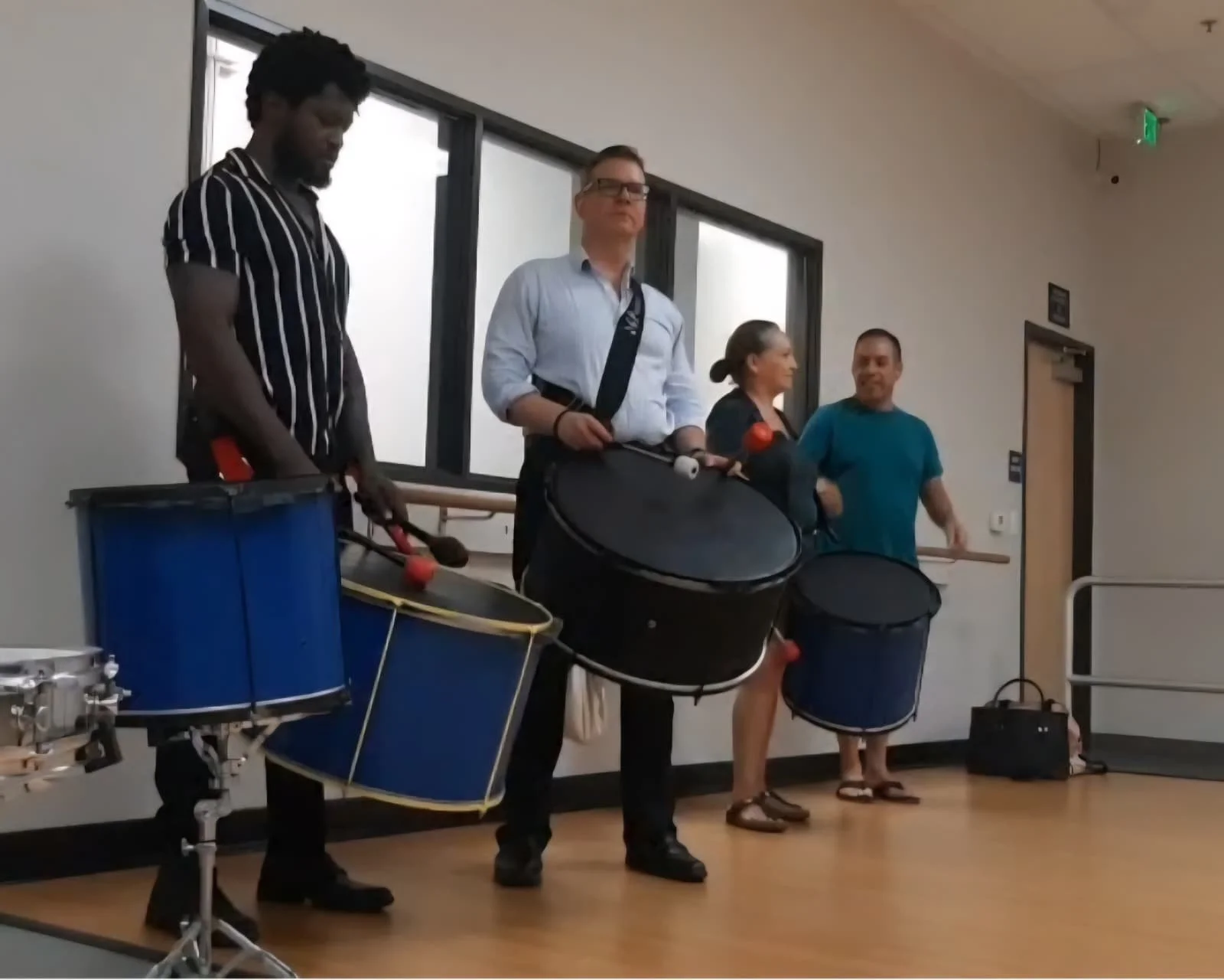 Four people standing on a stage playing drums with drumsticks, two women and two men, in a room with large windows and neutral walls. sacramento brazilian drum class