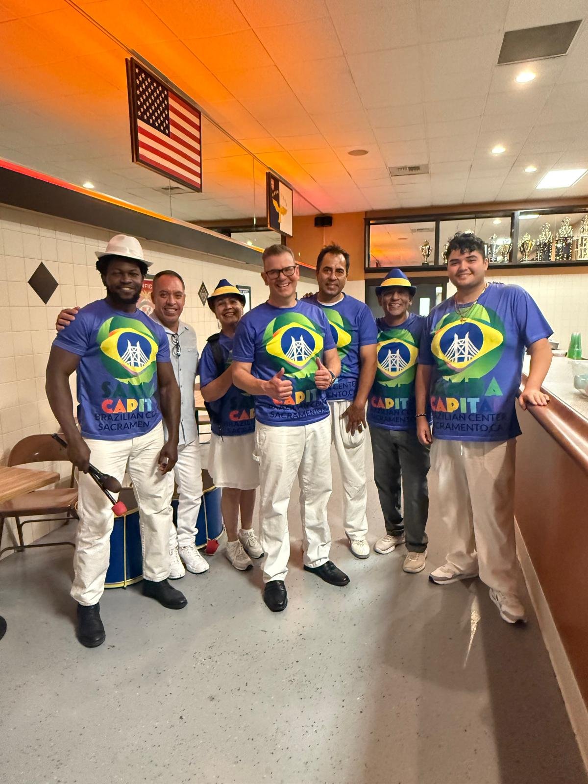Group of seven people wearing matching blue T-shirts with a bridge and Brazil flag logo, posing indoors with some smiling and giving thumbs up.  G.R.E.S. unidos da capital of sacramento brazilian samba performing arts group