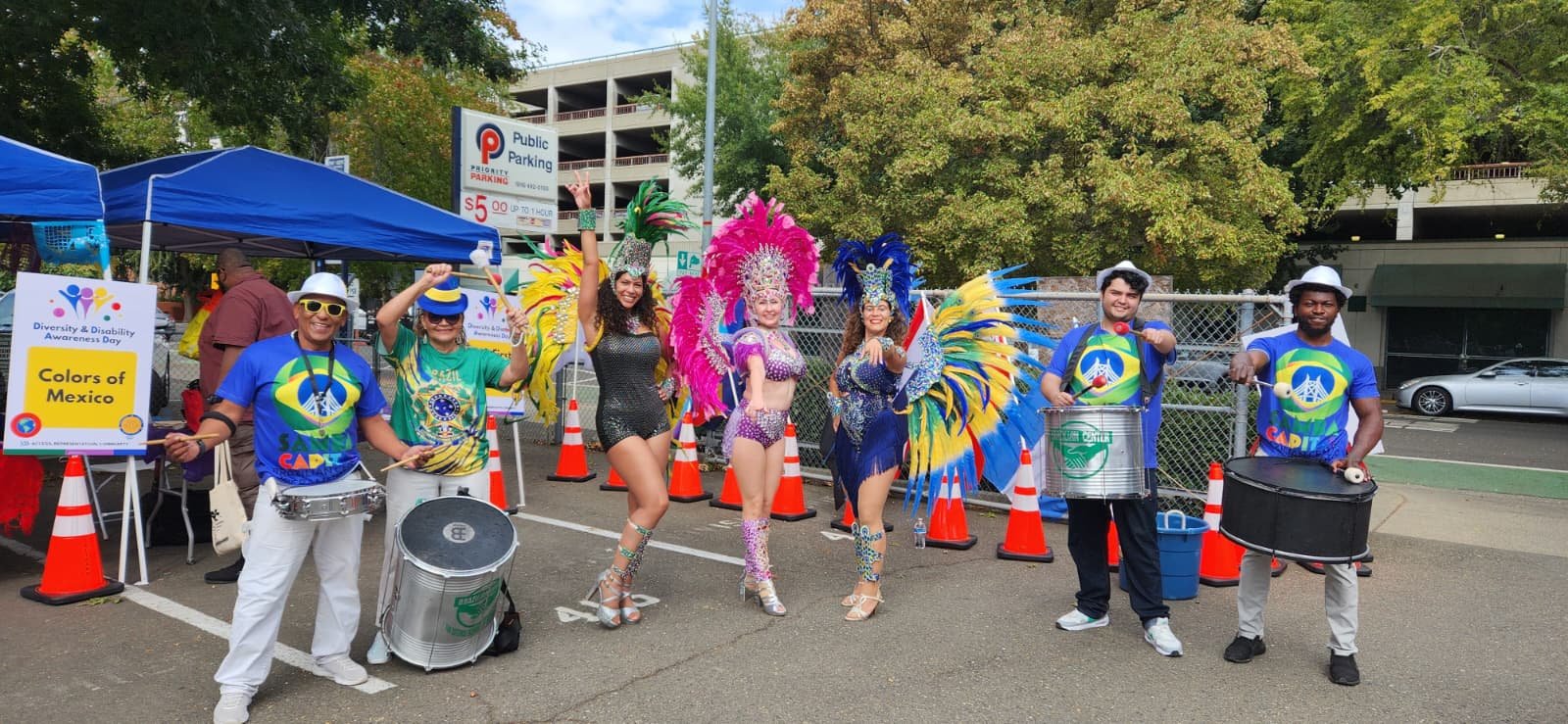 Group of parade performers in colorful costumes, some wearing feathered headdresses, standing in a parking lot with signs and orange cones around them.