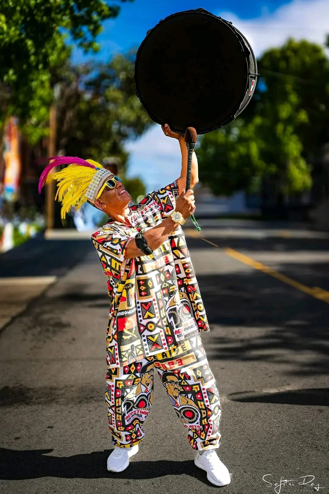 A man dressed in vibrant, patterned clothing and wearing a large yellow feathered headdress holds a large drum high above his head while standing on a street.