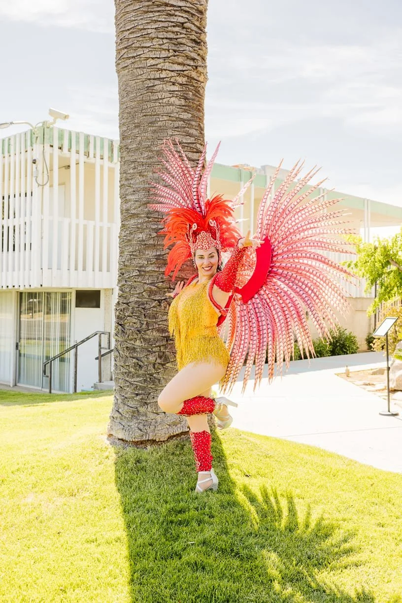 A woman in a colorful carnival costume with red feathers and yellow fringe, standing next to a palm tree on a grassy area, smiling and striking a pose.