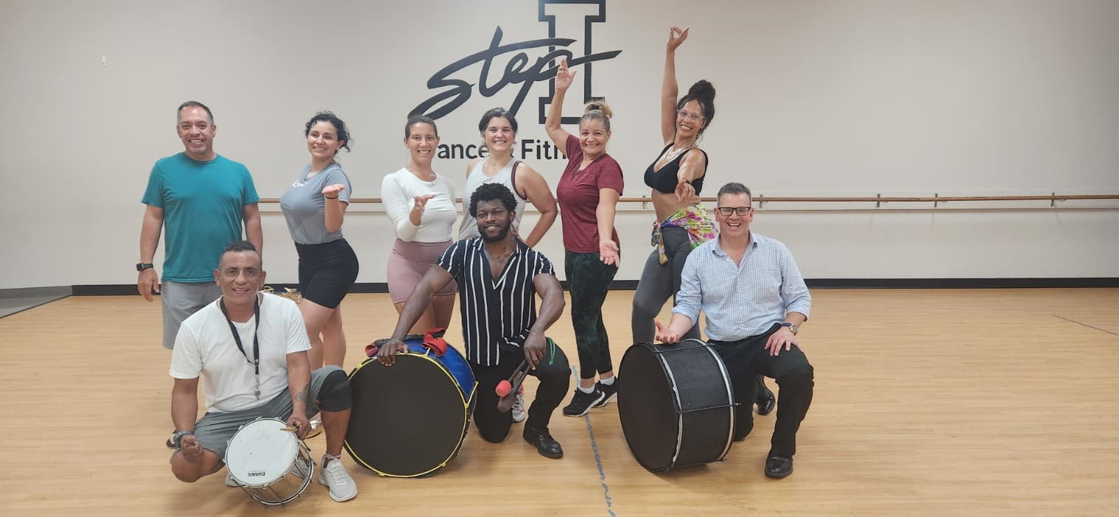 Group of nine people in a dance studio, smiling and posing for a photo. Some are holding musical instruments. Wall sign reads 'Step-1 Dance & Fitness.' Sacramento brazilian drum and samba class
