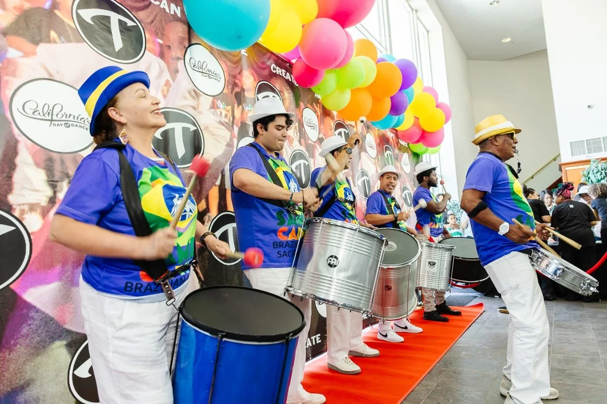 A group of musicians playing drums at a celebration event decorated with colorful balloons and a branded backdrop.  G.R.E.S. unidos da capital of sacramento brazilian samba performing arts group