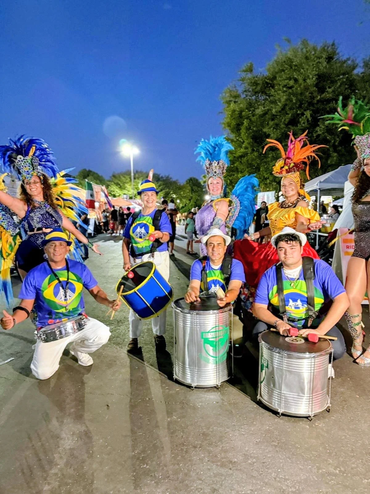 Group of people in colorful costumes and feathered headdresses, posing with drums and musical instruments at an outdoor event during the evening.