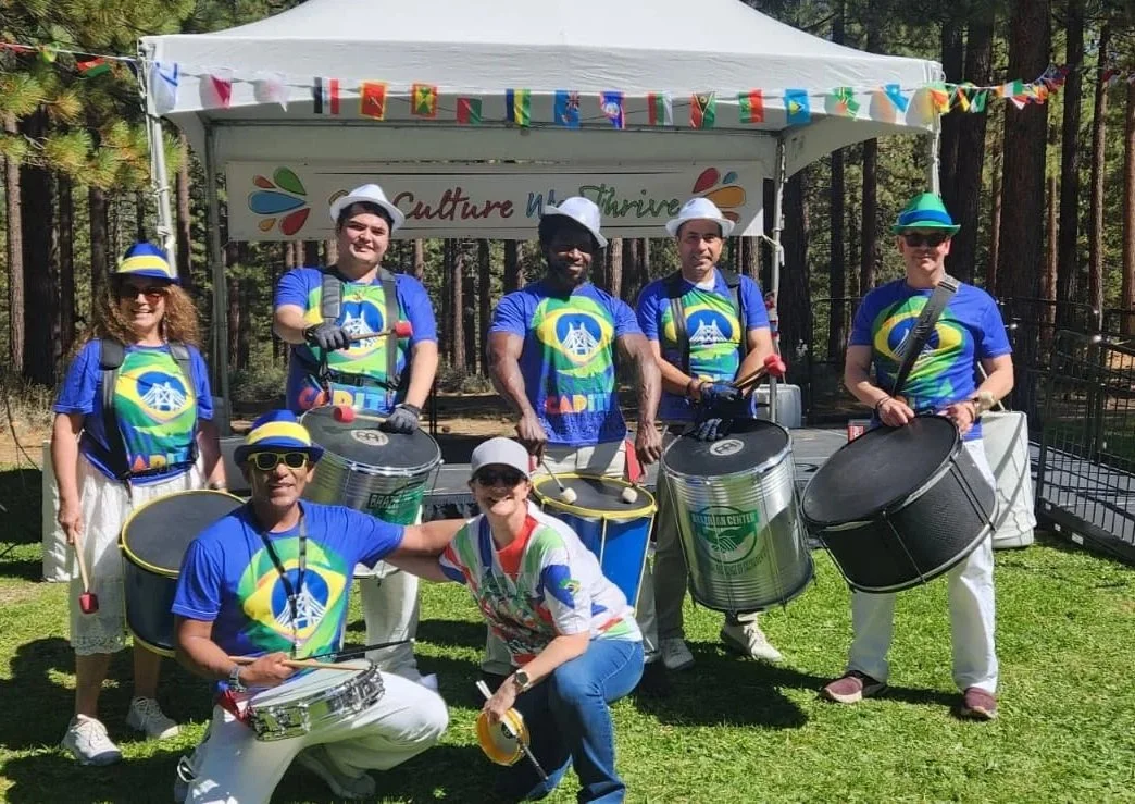 A group of seven people dressed in blue shirts with a green and yellow design, some wearing hats and sunglasses, posing with percussion instruments including drums and tambourines outdoors in front of a canopy with colorful bunting.