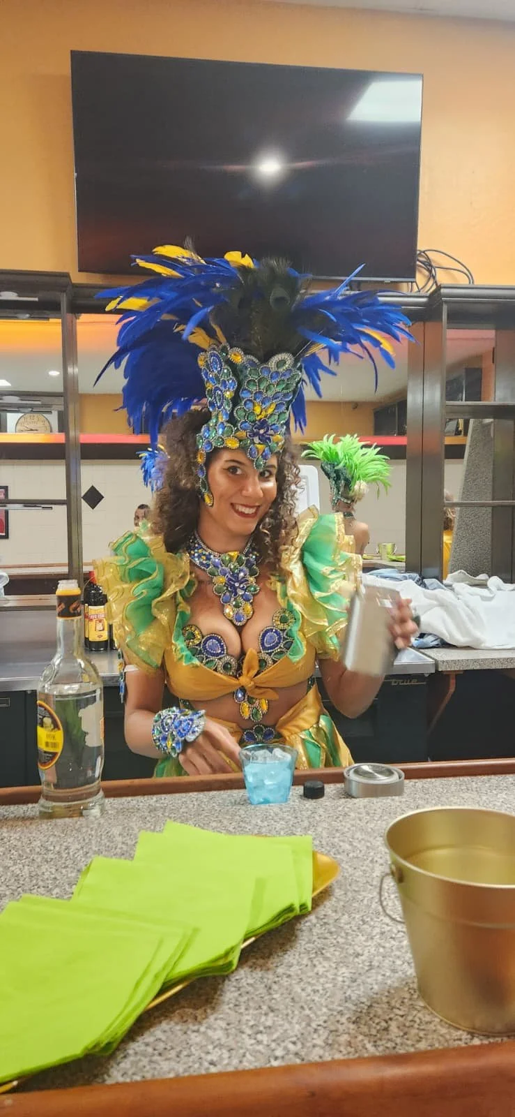 Woman in colorful carnival costume with blue, yellow, and green feathers and jewelry, holding a drink in a bar or restaurant setting.