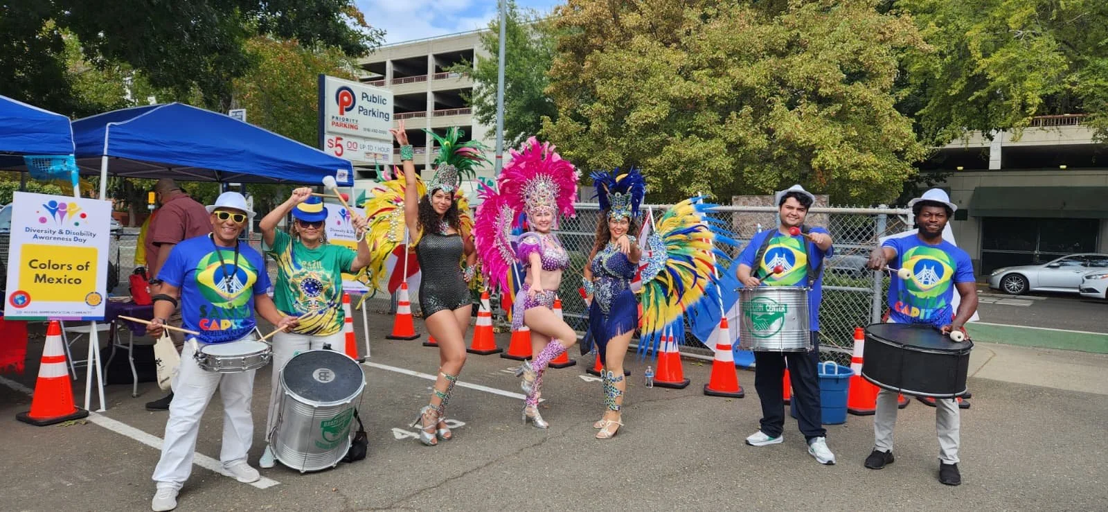 People dressed in colorful costumes participating in a parade for Diversity & Disability Awareness Day, with musical instruments, costumes with feathers, and a sign that says "Colors of Mexico".
