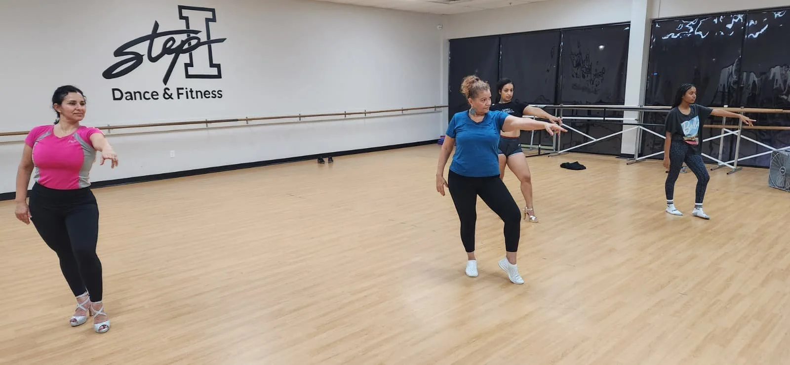 Four women are dancing in a dance and fitness studio with wooden floors and a wall marked 'Stop Dance & Fitness.' The women are dressed in athletic wear, and some are holding or extending their arms, suggesting they are practicing dance moves.