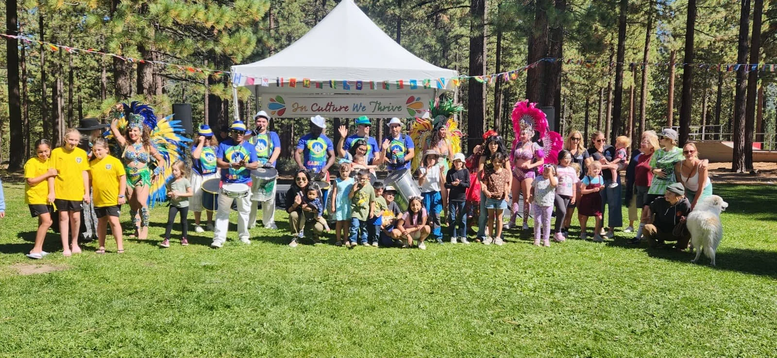 A group of children and adults gathered outdoors in front of a white tent decorated with colorful banners and a sign that reads 'The Culture We Thrive'. Some children are in colorful costumes, and there are performers in vibrant costumes with feather