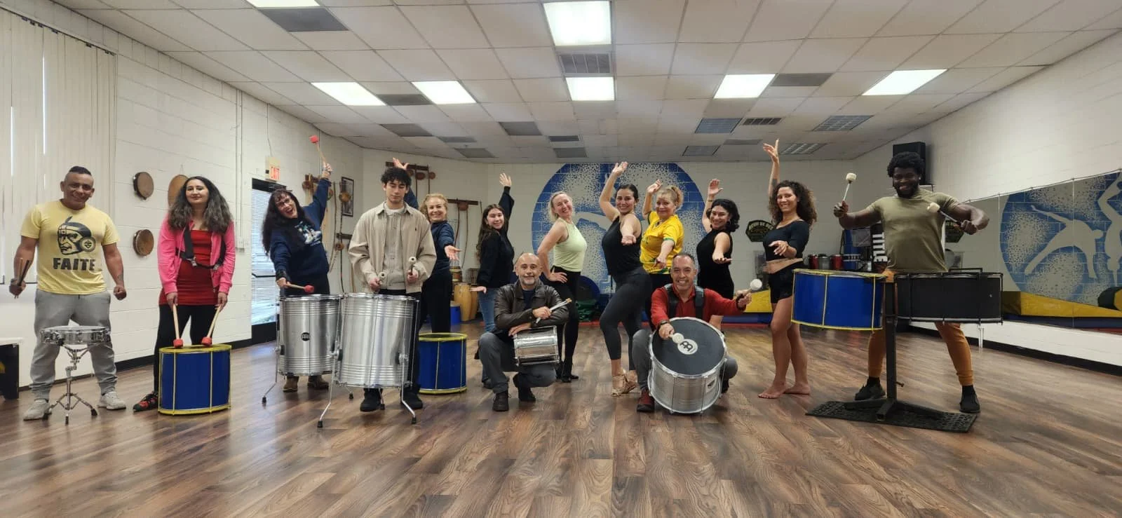 Group of people in a music rehearsal space holding percussion instruments and posing for a photo.  G.R.E.S. unidos da capital of sacramento brazilian samba performing arts group class